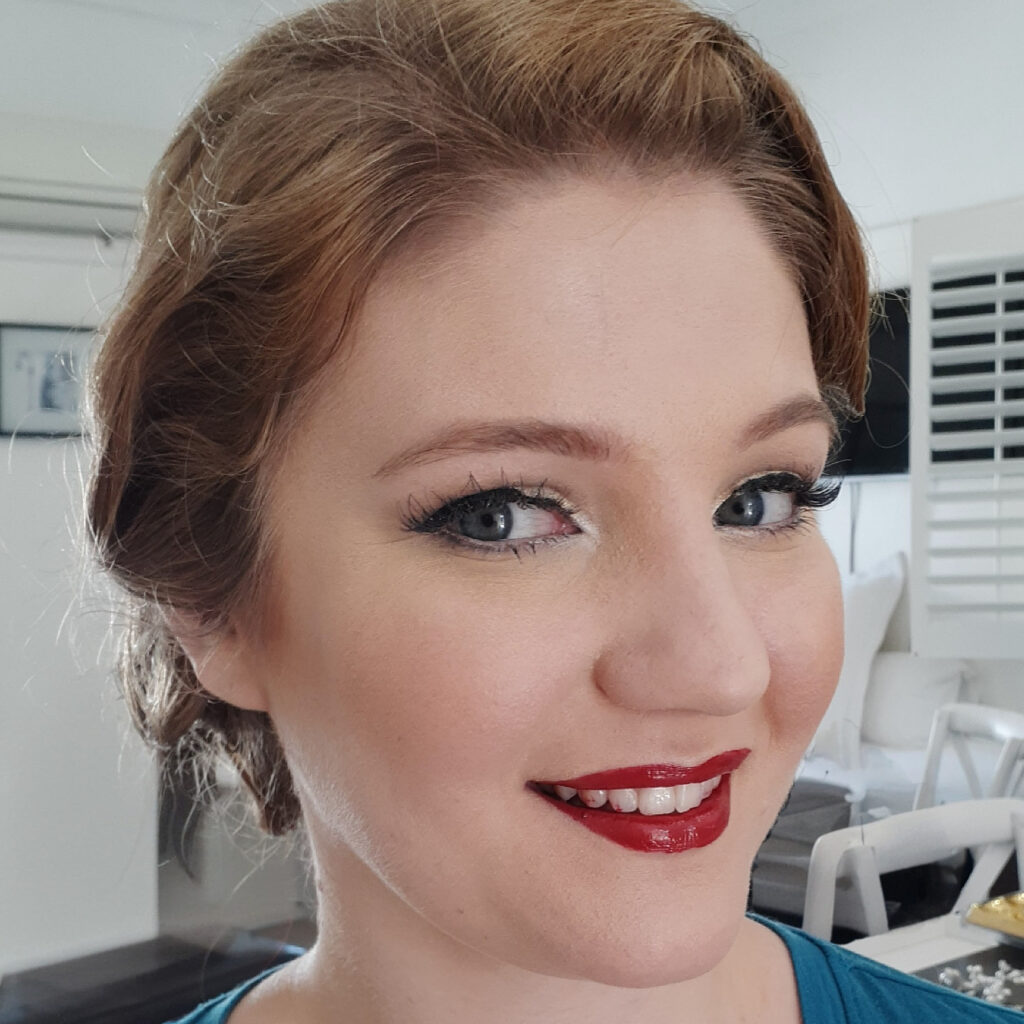 Close-up of a woman with auburn hair wearing bold red lipstick and defined eye makeup.