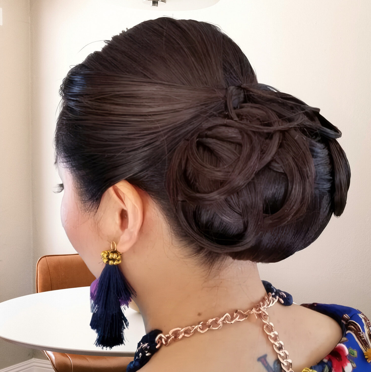 Dark hair styled in an intricate braided low bun, accessorized with navy tassel earrings and a gold chain necklace.