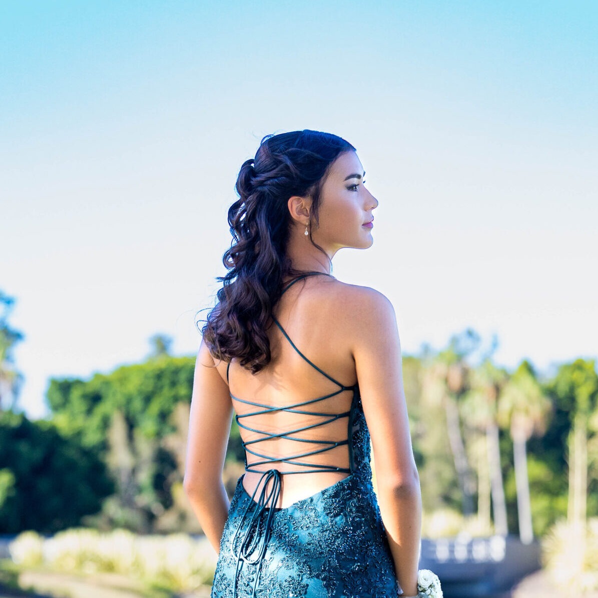 Young woman with curled half-up hairstyle wearing a teal lace dress with a lace-up back outdoors.