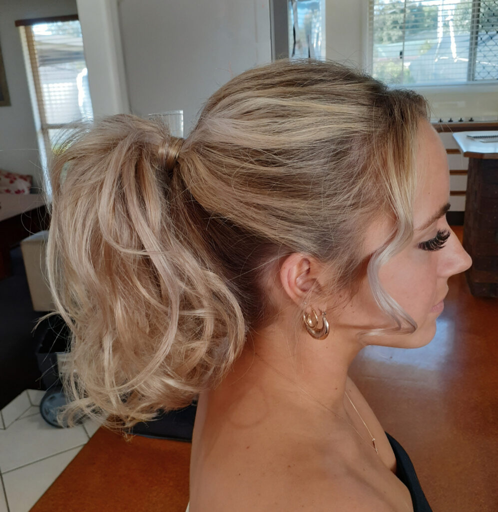 Woman with blonde hair styled in a textured high ponytail with loose strands framing her face.